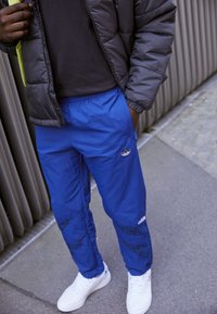 Pantaloni jogger blu con vita elasticizzata, dotati di una patch con logo e dettagli neri. Indossati con un giubbotto imbottito nero e sneakers bianche.
