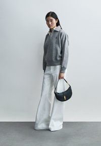 Γκρι πουλόβερ με φερμουάρ και ribbed μανσέτες, συνδυασμένο με λευκό παντελόνι φαρδύ. Το μοντέλο κρατάει μια μαύρη δερμάτινη τσάντα με χρυσές λεπτομέρειες.