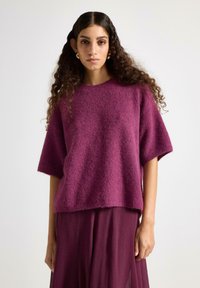 Maglione oversize fucsia con maniche corte, caratterizzato da un morbido tessuto lavorato a maglia. Abbinato a una gonna plissettata coordinata per un look armonioso.