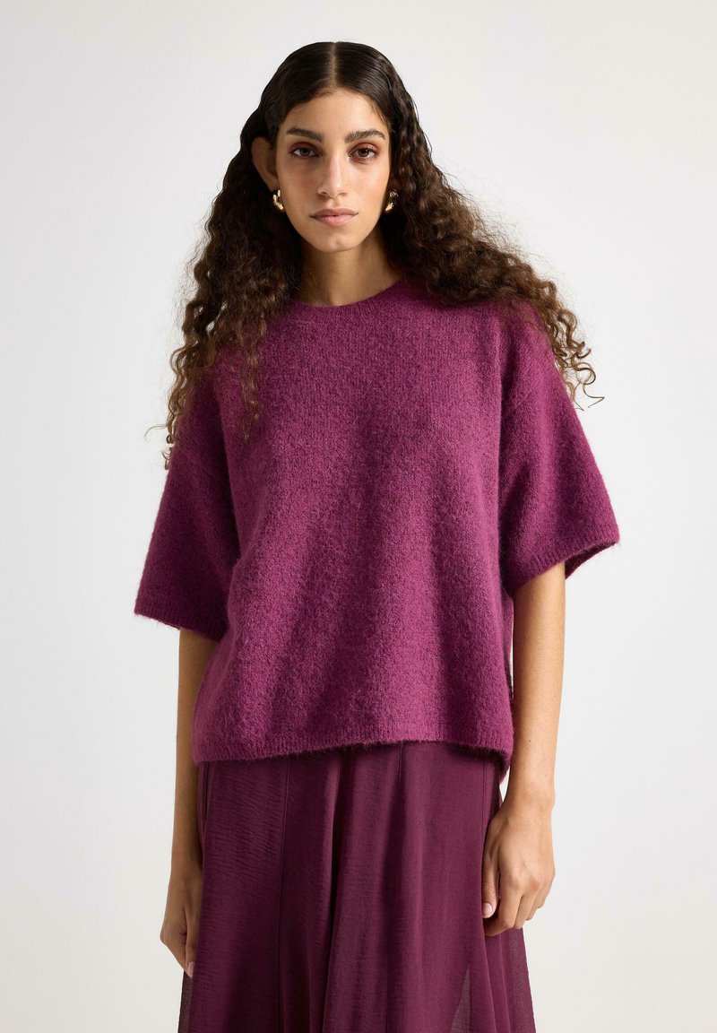 Maglione oversize fucsia con maniche corte, caratterizzato da un morbido tessuto lavorato a maglia. Abbinato a una gonna plissettata coordinata per un look armonioso.