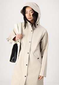 Beige vattentät jacka med huva, framsnappar och sidofickor; matchad med en svart väska med en fluffig vit kant.