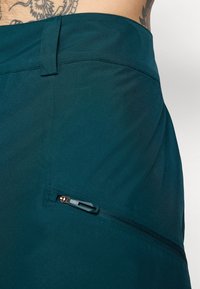 Smaragdgröna tygshorts med en slät yta, platt midjeband och en ficka med dragkedja på sidan med en svart dragflik.