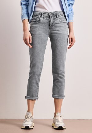 Person iført opsmøgede grå jeans, hvide sneakers, en hvid skjorte og en lyseblå jakke, der står mod en ensfarvet baggrund.