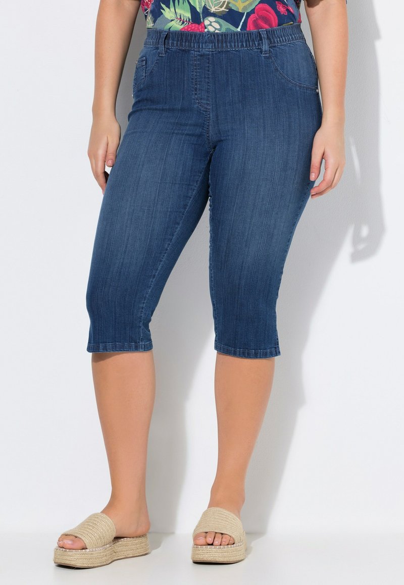 Tummansinisiä farkku-capri-housuja, joissa on joustava vyötärönauha, kaksi sivutaskua ja lahkeiden päättämät. Yhdistetty vaaleanvärisiin liukuhalkaisu-kengiin.