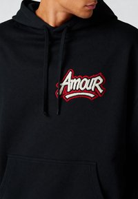 Felpa nera con cappuccio e tasca frontale, caratterizzata da un logo ricamato in rilievo rosso e bianco con la scritta "Amour" sul petto. Tessuto morbido e cordini regolabili.
