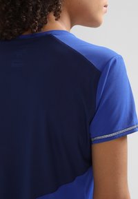 Blå sporttröja med texturerad tyg, en kontrasterande mörkblå panel på baksidan, korta ärmar och reflekterande accentdetaljer.