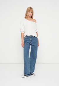 Biała, dziana sweter off-shoulder z bufiastymi rękawami, w połączeniu z niebieskimi spodniami typu wide-leg i czarnymi sneakersami. Minimalistyczny design, luźny krój.