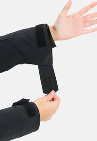 Black jacket cuff with Velcro fastener. Showing a hand lifting adjustable cuff, featuring fabric texture and sleek design. No visible patterns.