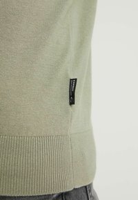 Pull en tricot vert clair avec ourlet côtelé et côtés côtelés, comportant une petite étiquette de marque noire sur le côté gauche près de la couture.