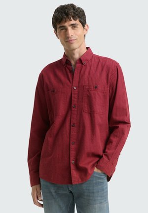 Chemise rouge à manches longues avec un col à bouton, deux poches avant et une texture à rayures subtile. Portée avec un jean bleu clair.
