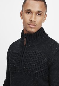 Sort strikket sweater med høj krave og kvart lynlås. Har strukturerede mønstre og farverige pletter i stoffet.
