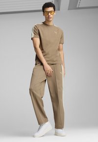 Tan T-shirt med lysebeige detaljer og Puma-logo; matchende tan sweatpants med hvide sidestriber; hvide sneakers; minimalistisk design.