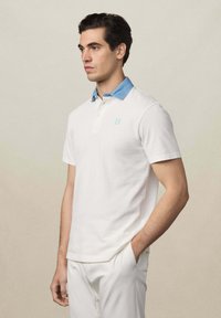 Weißes Poloshirt mit einem hellblauen Denim-Kragen und einem kleinen blauen Logo. Kurzärmlig und aus weichem Baumwollmaterial. Sauberes, einfaches Design.