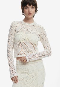Top lungo bianco a maniche lunghe, tagliato, realizzato in pizzo intricato, con un motivo a onda e texture a coste, abbinato a una gonna coordinata.