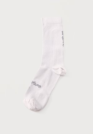 Une chaussette blanche mi-mollet avec des panneaux en maille texturée et un texte noir près des orteils et le long du côté, posée à plat sur un fond clair.