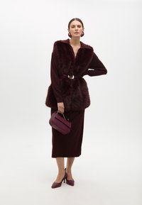 Burgundské dlouhé šaty s dlouhým rukávem a umělou kožešinou, stahované v pase. Doplňky zahrnují malou kabelku a lodičky s úzkou špičkou.