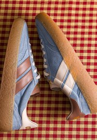 Paire de baskets en daim bleu avec des bandes en cuir marron et blanc, des lacets blancs et des semelles en gomme texturée sur un tissu à carreaux rouge et jaune.