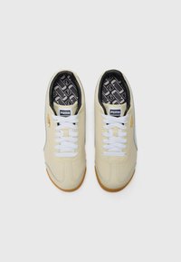 Light beige sneakers with white laces and a black patterned insole. Features gray accents and a gum rubber sole. Brand logo on tongue.