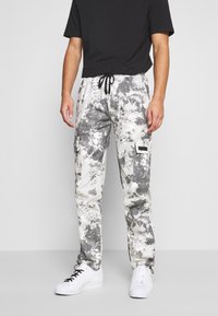 Mann in schwarzem T-Shirt, grauen und weißen Camouflage-Cargo-Hosen und weißen Sneakers, der vor einem schlichten Hintergrund steht.