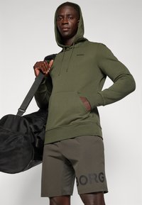 Felpa verde con cappuccio e cordino, abbinata a pantaloni corti coordinati con logo "BORG" nero. Borsa da palestra nera con tracolla inclusa.