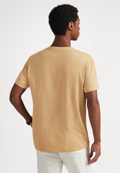 Īsām piedurknēm, apaļā kakla t-krekls gaiši brūnā krāsā, izgatavots no mīkstas kokvilnas. Gluda tekstūra ar vaļēju piegriezumu, ar minimālām šuvēm.