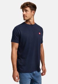 Camiseta de algodón azul marino con cuello redondo y mangas cortas, que presenta un logo rojo en el lado izquierdo del pecho. Textura suave y corte clásico.