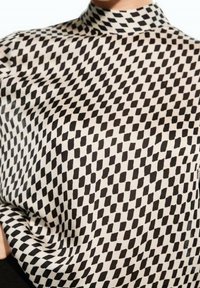 Schwarz-weiß gemusterte Bluse mit hohem Kragen. Der Stoff weist ein sich wiederholendes Diamantmuster sowie eine weiche, glatte Textur auf.