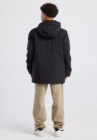 Musta vedenpitävä takki, jossa on huppu ja yksinkertainen muotoilu, päällä vaaleanruskeat housut ja valkoiset lenkkarit mustilla yksityiskohdilla.