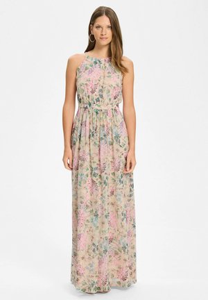 Junge Frau steht frontal und trägt ein ärmelloses, bodenlanges Kleid mit floralen Mustern in Pastellrosa, Blau und Grün auf beigem Hintergrund.