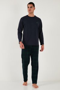 Man die een marineblauw shirt met lange mouwen draagt met een hert-logo en groene en marineblauwe geruite pyjamabroek, staand op blote voeten tegen een effen witte achtergrond.