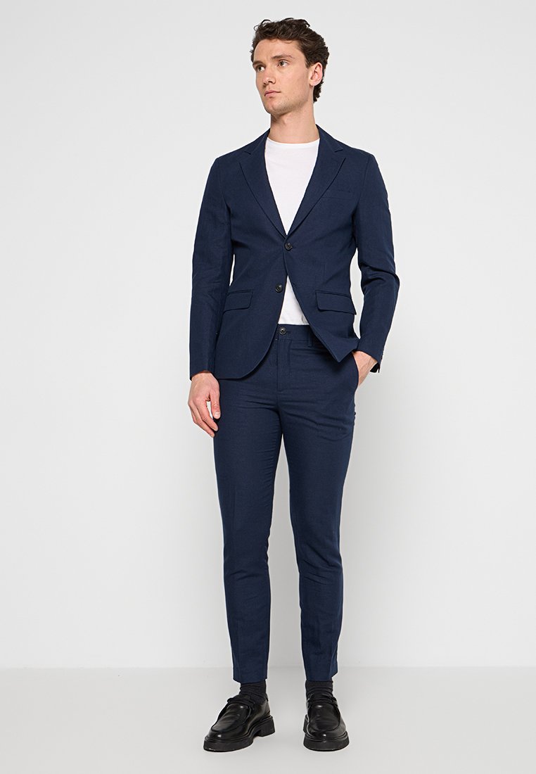 Marineblauer maßgeschneiderter Anzug mit einem einreihigen Blazer mit Reverskragen, zwei Vordertaschen und schlank geschnittenen Hosen. Trägt ein weißes T-Shirt darunter.
