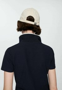 Marineblå polo T-shirt med hvid krave; båret bagfra. Lys beige baseballcap på kort, bølget hår. Enkel design, afslappet look.