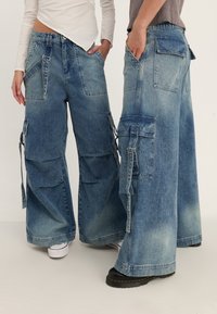 Blå denim cargo-byxor med vida ben, med flera sidofickor och en blekt finish. Designen inkluderar en blandning av texturer och sömmar.