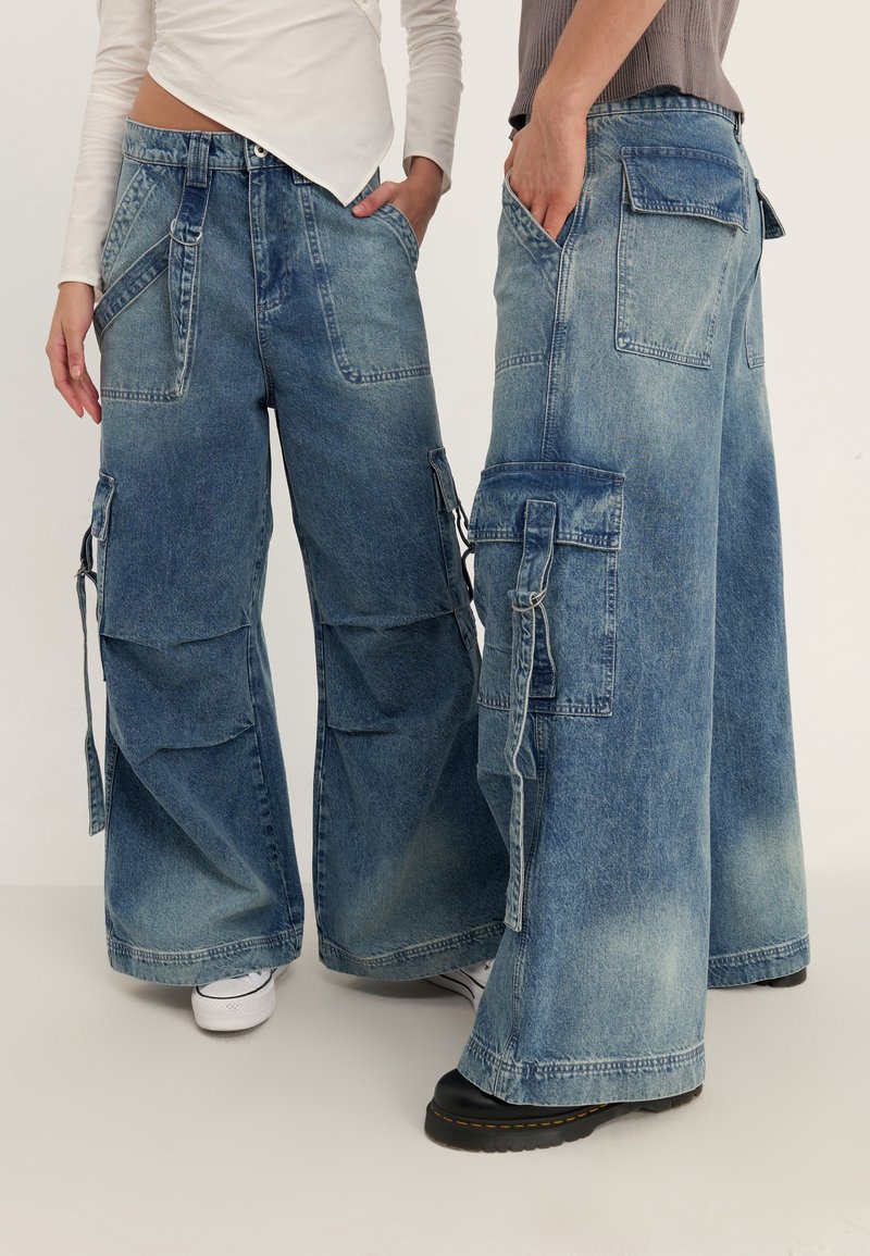 Blå denim cargo-byxor med vida ben, med flera sidofickor och en blekt finish. Designen inkluderar en blandning av texturer och sömmar.