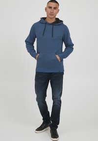 Blå hættebluse med forlommer, snoretræk og ribbede manchetter, parret med mørke jeans og sorte sneakers.