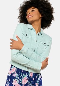 Kerge piparmündiroheline lühikeste varrukatega denim-jakk, millel on nupud ja rinna taskud, kombineeritud lillelise tumesinise seelikuga. Pehme tekstuur.