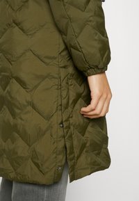 Chaqueta acolchada verde con un acabado texturizado, que presenta una abertura lateral y un cierre de botones a la vista. La manga tiene un puño elástico para un mejor ajuste.