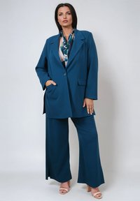 Teal oversized suit featuring a single-button blazer and wide-leg trousers. The fabric is smooth with a slight sheen. Long sleeves and pockets.