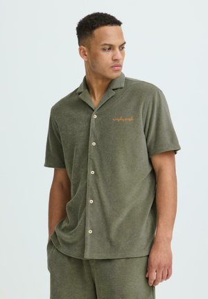 Homme portant une chemise à manches courtes olive avec boutons et un short assorti, regardant sur le côté les mains dans les poches.