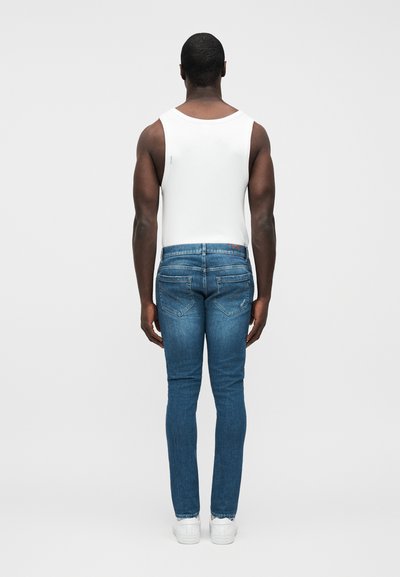Jean bleu slim avec une taille mi-haute, associé à un débardeur blanc ajusté. Le jean présente des poches arrière et une texture denim classique.