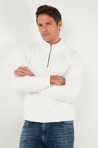 Valge tõmbluku ja kõrge kraega sviiter, millel on pikad varrukad ja sile tekstuur. Kantud siniste teksadega, käed risti.