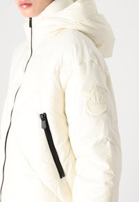 Krēma krāsas pūkains žakets ar kapuci, melnu rāvējslēdzēju un gumijas logo uz piedurknes. Raksturo izšūta tekstūra un elegants dizains.