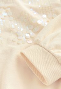 Světle béžový materiál mikiny s žebrovaným manžetem. Obsahuje duhový flitrový vzor uspořádaný vertikálně na látce.