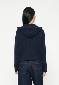 Granatowy krótki hoodie o prążkowanej strukturze i z kapturem. Połączony z ciemnoniebieskimi dżinsami z dwoma tylnymi kieszeniami i metką z logo.