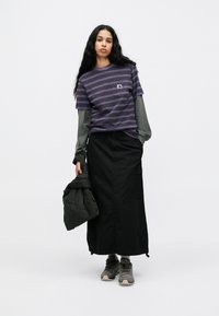 Vijolična majica s črtami, preletena čez sivo dolg rokav, v kombinaciji z črno krilom do gležnjev in sivimi športnimi copati; drži črno puhovko.