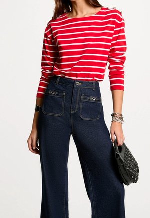 Mujer con camisa de manga larga a rayas rojas y blancas, vaqueros de cintura alta azul oscuro con bolsillos delanteros, sosteniendo un bolso negro con tachuelas.