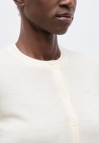 Prim-plan cu o persoană care poartă un cardigan cu nasturi, de culoare alb-crem, cu decolteu rotund, pe un fundal simplu.