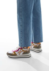 Sneakers con un design bianco, maculato e in oro metallizzato. Presentano lacci rosa e una suola nera spessa. Indossati con jeans azzurri.