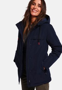 Veste d'extérieur bleu marine avec capuche, dotée d'une fermeture éclair et de boutons, de deux poches avant, et de cordons de serrage noirs avec des accents rouges.