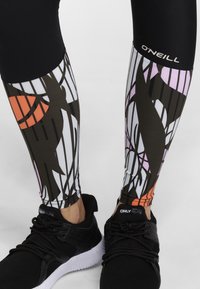 Leggings neri con un motivo astratto che presenta forme arancioni, rosa e marroni, abbinati a scarpe da ginnastica nere con suole bianche.
